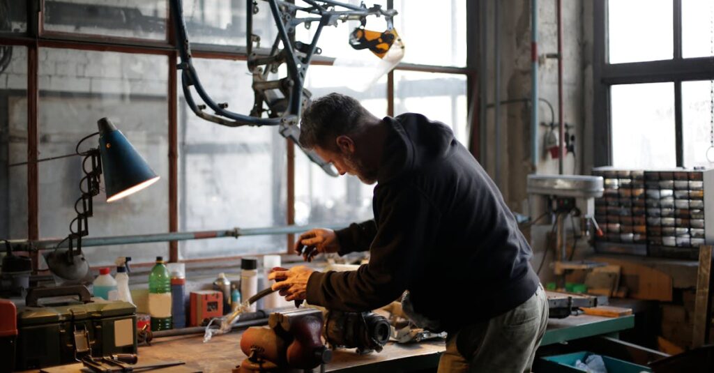 Back view of unrecognizable male mechanic in workwear standing near workbench while fixing broken metal detail in lamplight in modern workshop